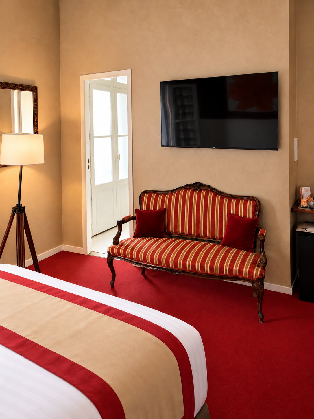 Chambre d'hôtel aux murs beiges avec une banquette ancienne à rayures rouges, un lit au chemin de lit rouge et beige, un téléviseur mural et un lampadaire.