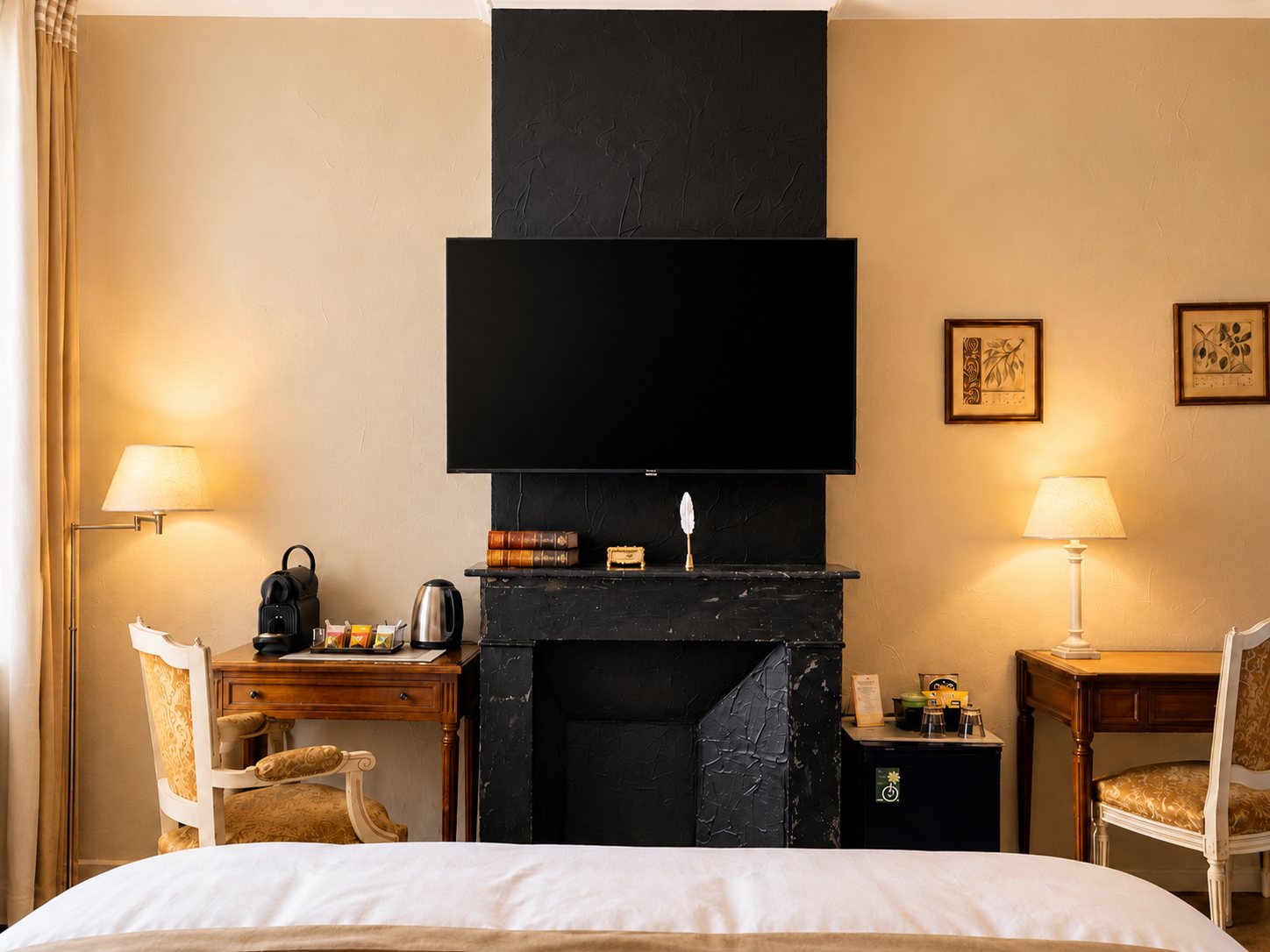 Chambre d'hôtel aux murs beige avec une cheminée en marbre noir surmontée d'une télévision, deux bureaux en bois, des fauteuils dorés et un coin café.