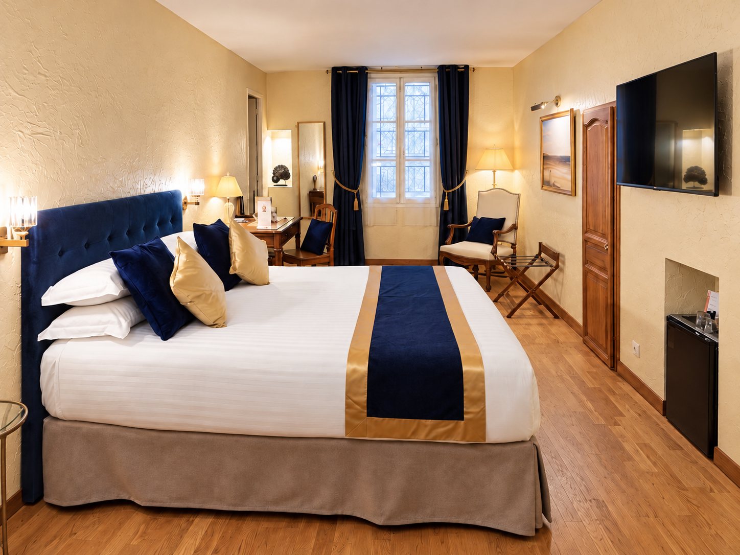 Chambre d'hôtel lumineuse avec un grand lit à tête de lit en velours bleu marine, un chemin de lit doré, un bureau en bois, un fauteuil et un téléviseur mural.