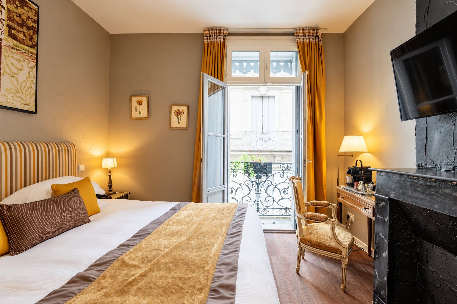 Chambre d'hôtel aux tons chauds avec un grand lit, des rideaux dorés, une porte-fenêtre ouverte sur un balcon en fer forgé, une cheminée en marbre noir et un fauteuil ancien.