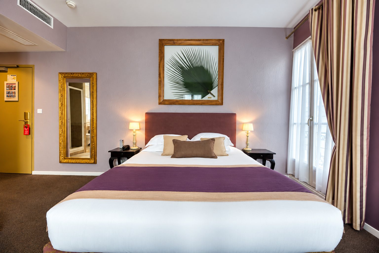 Chambre d'hôtel aux murs mauves avec un grand lit à plaid violet, deux lampes de chevet, un miroir doré et de longs rideaux rayés devant la fenêtre.