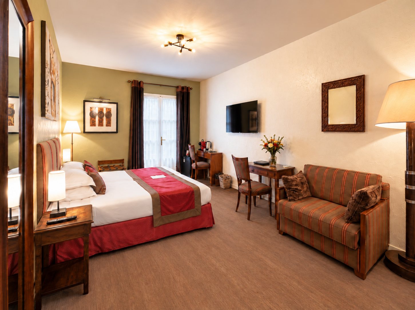 Chambre d'hôtel aux tons chauds avec un grand lit à chemin de lit rouge, un canapé rayé, un bureau en bois et une télévision murale.