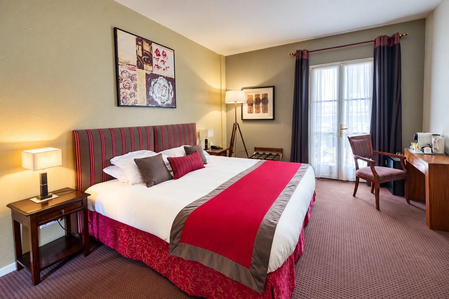 Chambre d'hôtel lumineuse avec un grand lit à chemin de lit rouge, deux tables de chevet, un bureau en bois et une porte-fenêtre aux rideaux sombres.