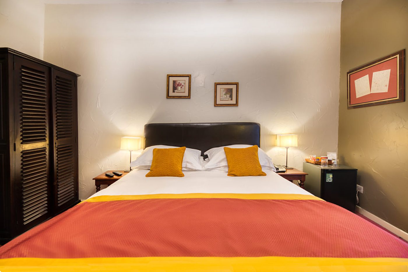 Chambre d'hôtel lumineuse avec un grand lit double habillé d'un plaid orange et de coussins jaunes, deux lampes de chevet et une armoire en bois sombre à persiennes.