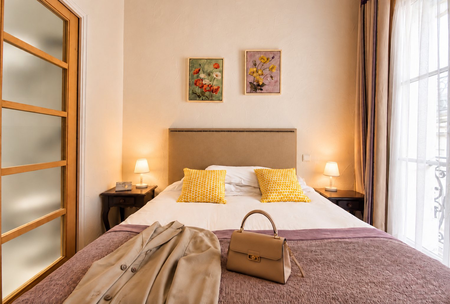 Chambre élégante avec un lit double orné de coussins jaunes, deux tableaux de fleurs au mur, des lampes de chevet allumées, un manteau beige et un sac à main posés sur le couvre-lit.