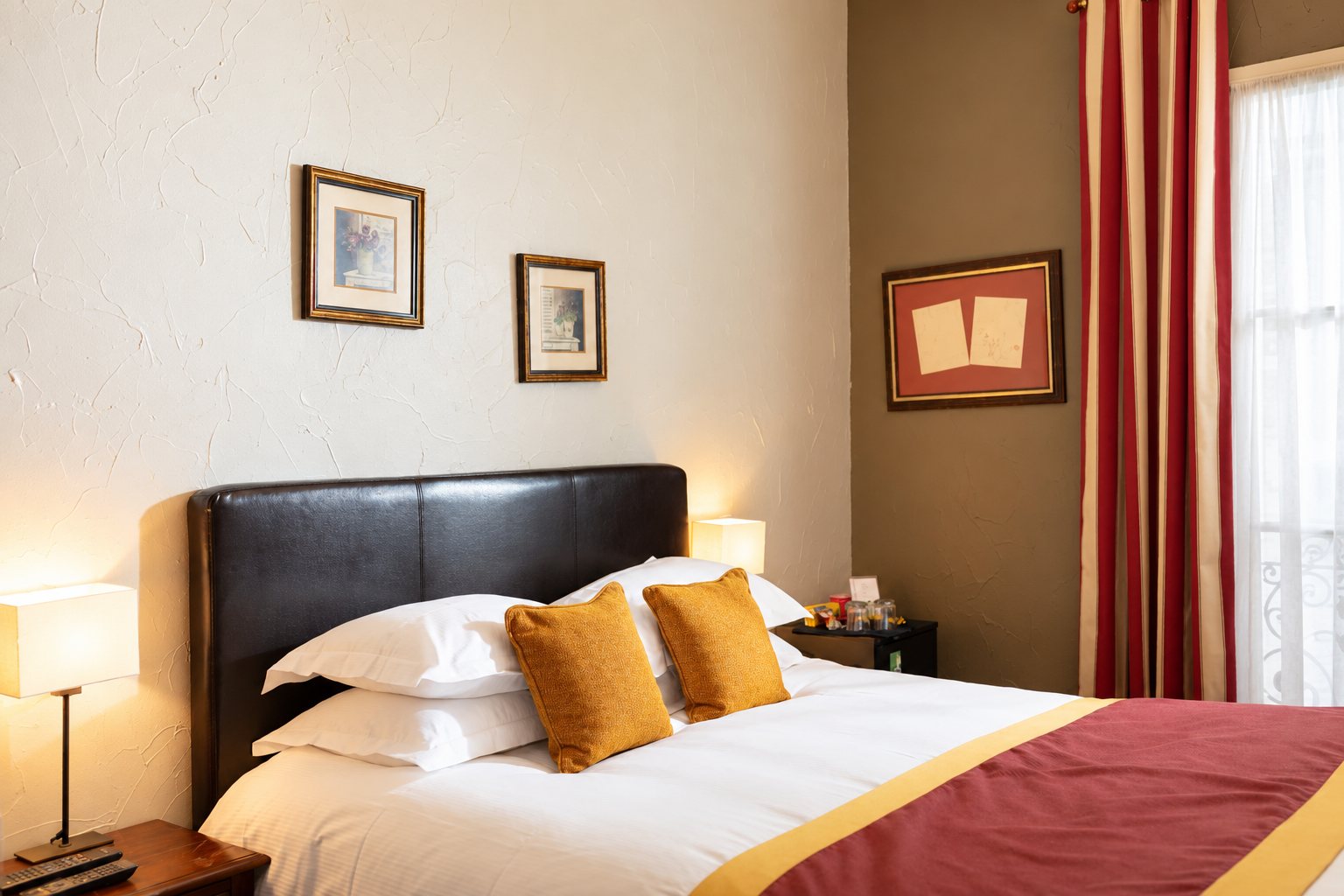 Chambre d'hôtel chaleureuse avec un lit double à tête de lit en cuir sombre, des coussins moutarde, des cadres au mur et des rideaux rayés rouge et crème.