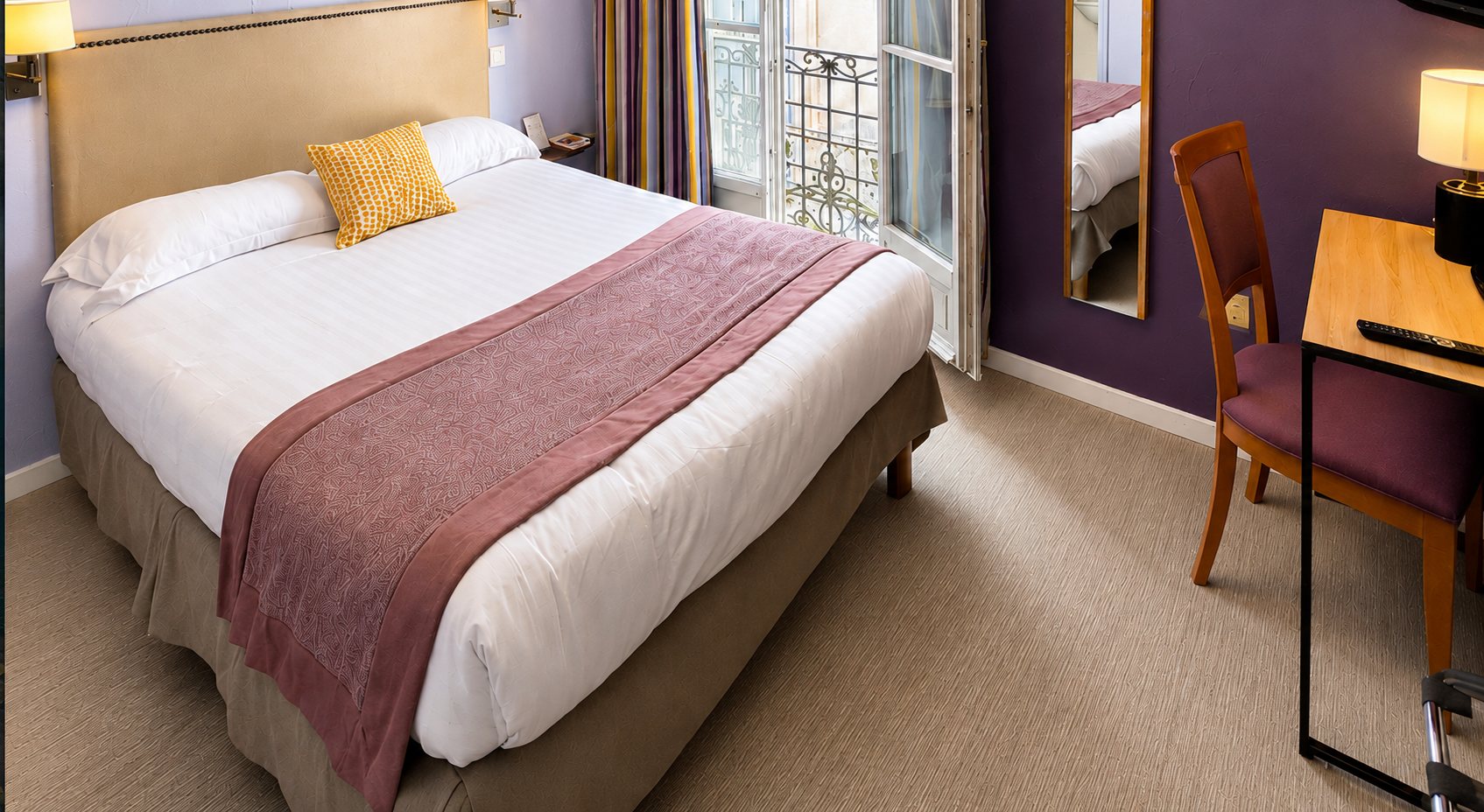 Chambre d'hôtel lumineuse avec un grand lit double habillé de blanc et d'un chemin de lit mauve, un bureau en bois, une chaise et une porte-fenêtre donnant sur un balcon en fer forgé.