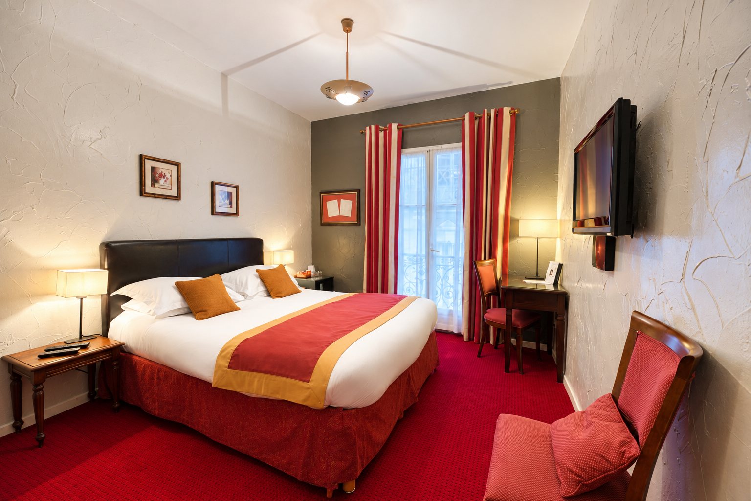 Chambre d'hôtel chaleureuse avec un lit double orné d'un chemin de lit rouge et orangé, une moquette rouge, un bureau, une télévision murale et une porte-fenêtre à rideaux rayés.