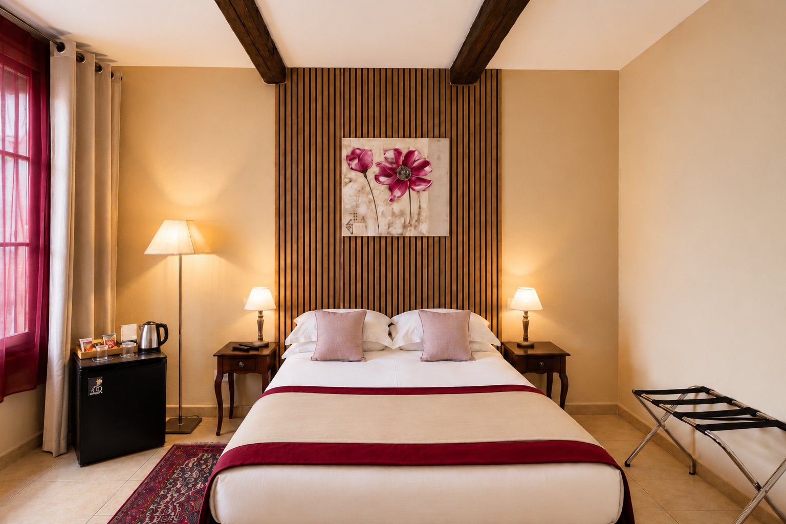 Chambre d'hôtel aux tons chauds avec un lit double, une tête de lit en tasseaux de bois ornée d'un tableau de fleurs roses, deux chevets avec lampes et un minibar.