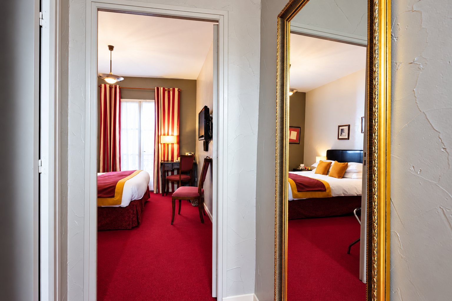 Une chambre d'hôtel aux rideaux rayés rouge et crème et à la moquette rouge, vue par une porte ouverte et reflétée dans un grand miroir au cadre doré.