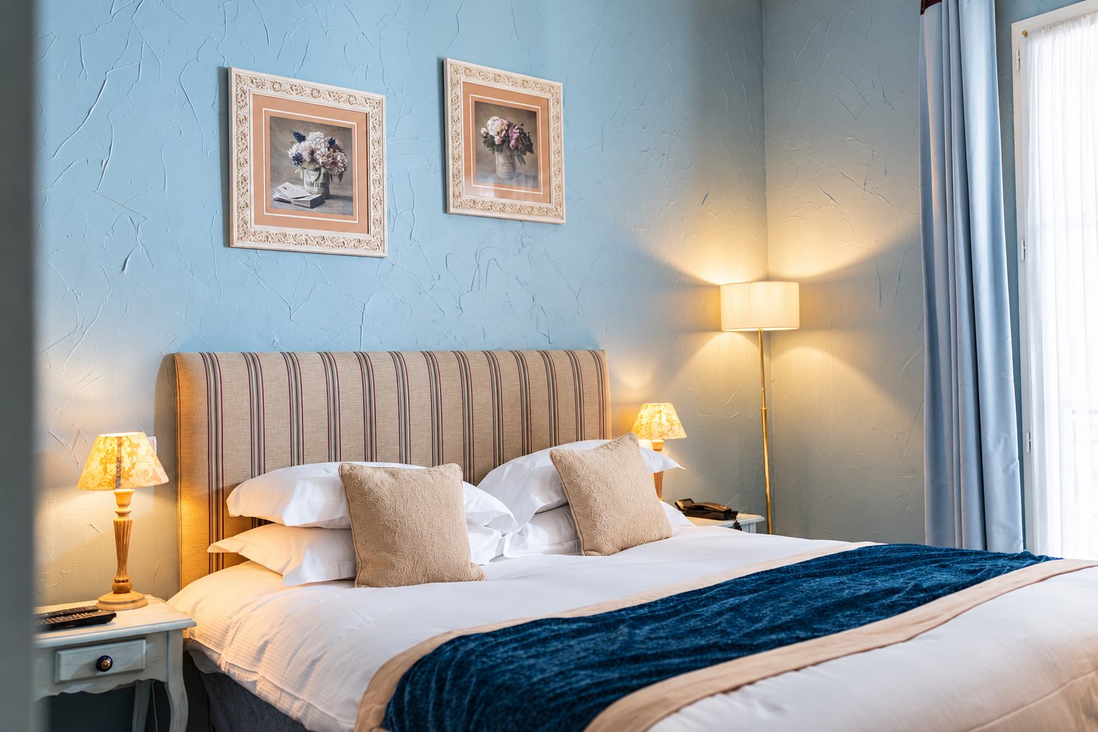 Chambre d'hôtel aux murs bleus texturés avec un grand lit double drapé de blanc, deux cadres floraux, des lampes de chevet allumées et un rideau bleu.