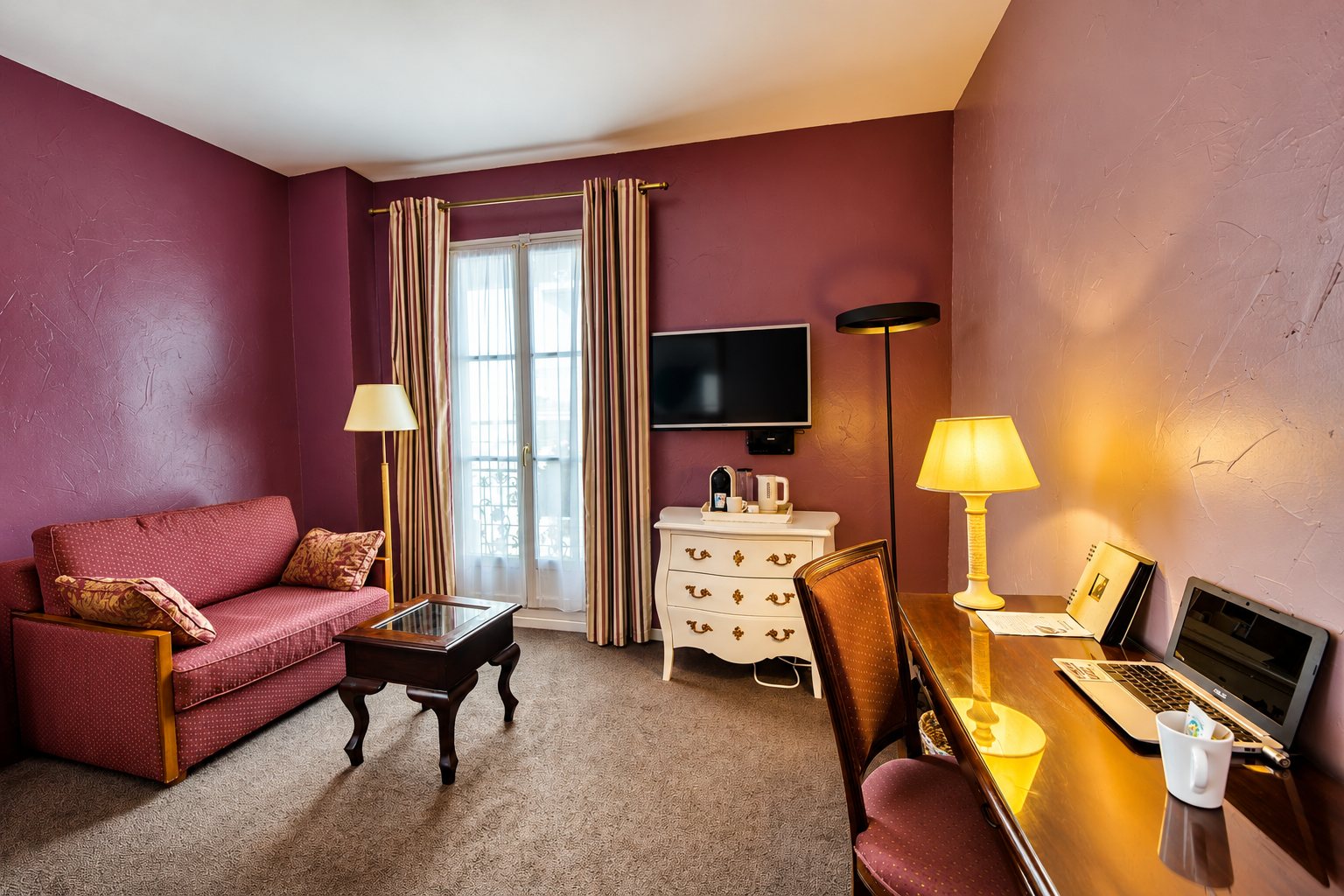 Chambre d'hôtel aux murs couleur prune, équipée d'un canapé rouge, d'une table basse, d'une commode blanche, d'un téléviseur et d'un bureau avec ordinateur portable.