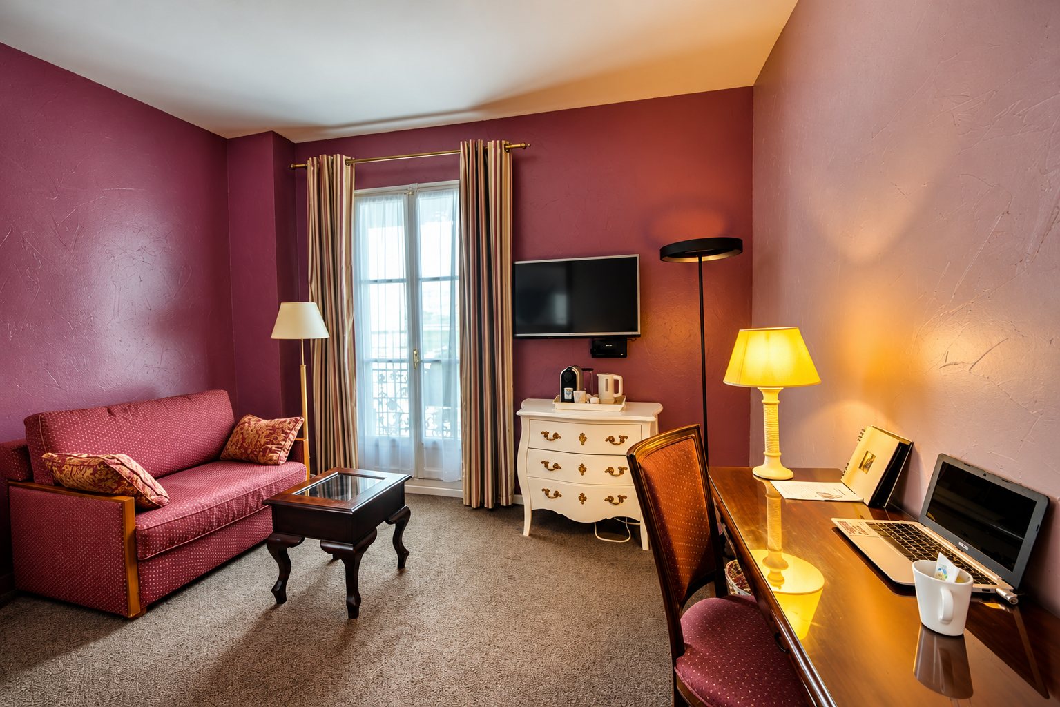 Chambre d'hôtel aux murs prune équipée d'un canapé rouge, d'une commode blanche, d'une télévision murale et d'un bureau avec ordinateur portable et lampe allumée.