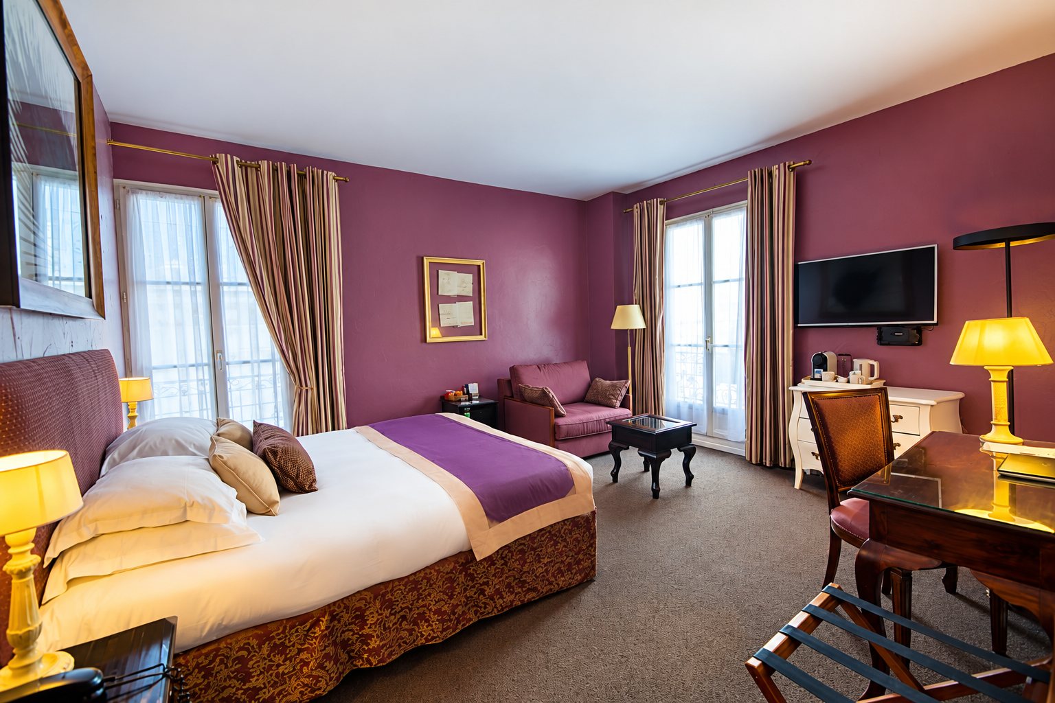 Chambre d'hôtel élégante aux murs violets, avec un grand lit, un fauteuil, un bureau, une télévision murale et de hautes fenêtres drapées de rideaux rayés.