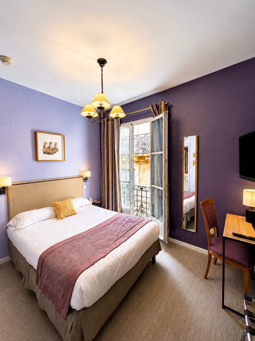 Chambre d'hôtel lumineuse aux murs violets, avec un lit double, un lustre à abat-jour jaunes, un bureau et une porte-fenêtre ouverte sur un balcon.