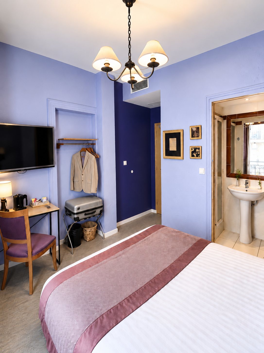 Chambre d'hôtel aux murs lavande et bleu foncé avec un lit double, un bureau, une valise sur son support, un lustre et une salle d'eau avec lavabo sur pied.