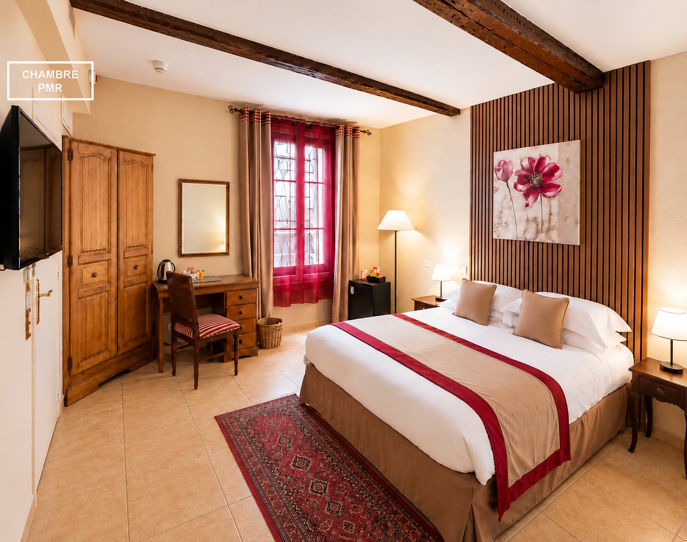 Chambre d'hôtel adaptée PMR avec grand lit double, armoire en bois, bureau, tapis rouge et mur de lattes en bois décoré d'un tableau de fleurs.