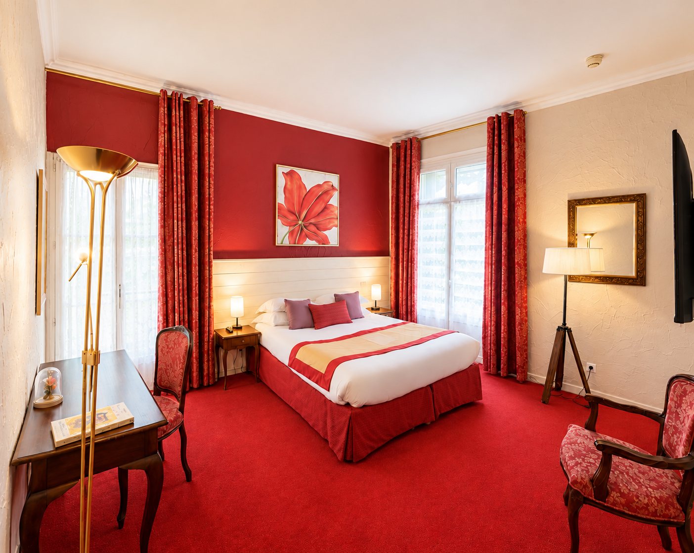 Chambre d'hôtel aux murs et à la moquette rouges, avec un lit double, des rideaux à motifs, un bureau en bois et des fauteuils de style.