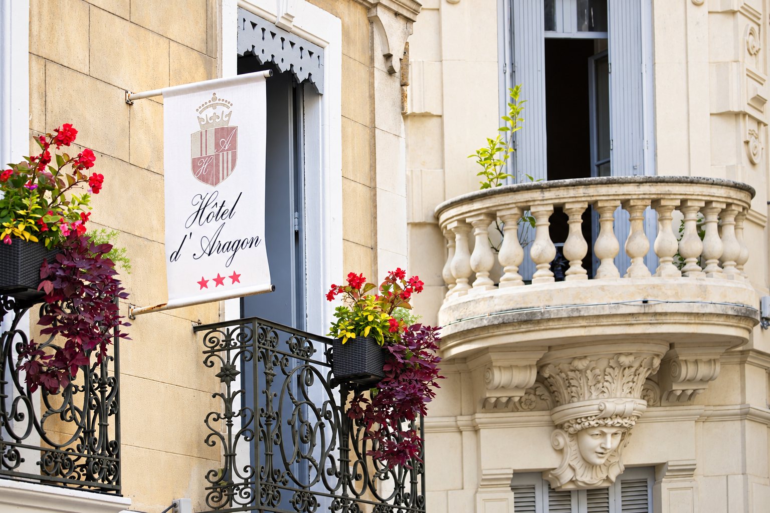 Une enseigne blanche de l'Hôtel d'Aragon, trois étoiles, est fixée à une façade en pierre ornée de balcons en fer forgé fleuris et d'un balcon sculpté.