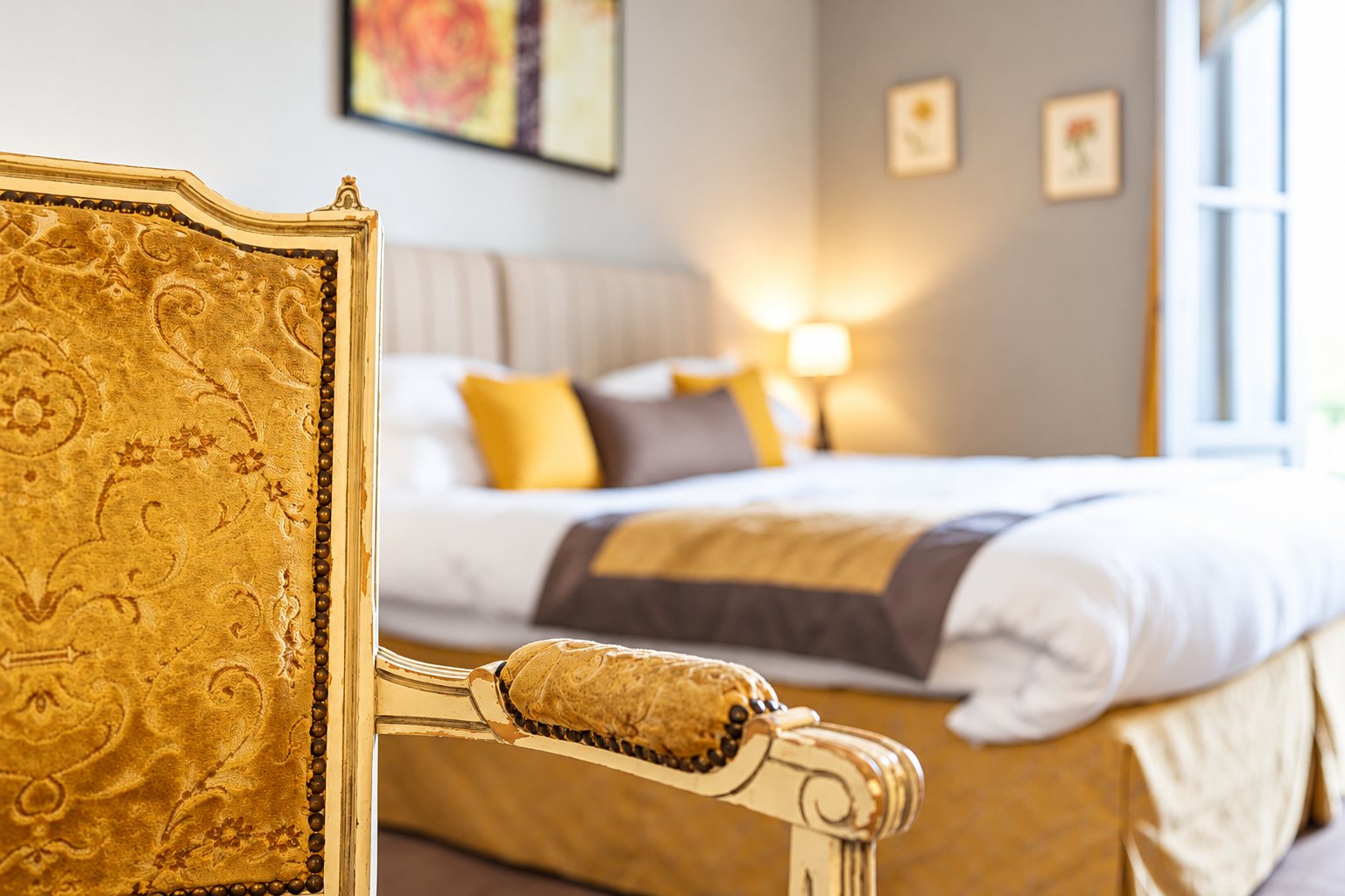 Gros plan sur un fauteuil de style Louis XVI en velours doré au premier plan, avec un lit aux coussins jaunes flou dans une chambre lumineuse.