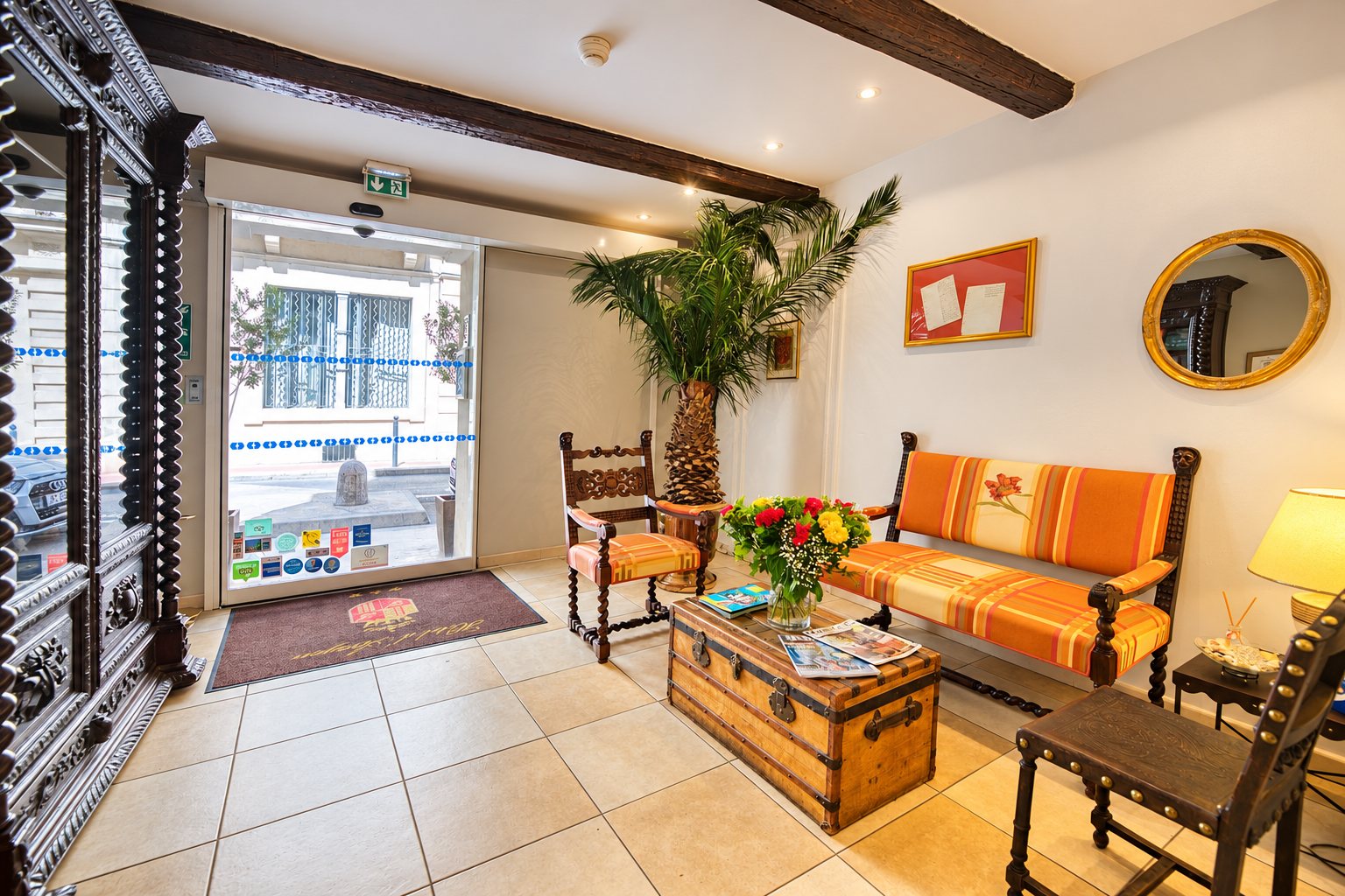 Hall d'entrée lumineux avec une banquette rayée orange, une vieille malle servant de table basse, un bouquet de fleurs, un palmier et une porte vitrée donnant sur la rue.