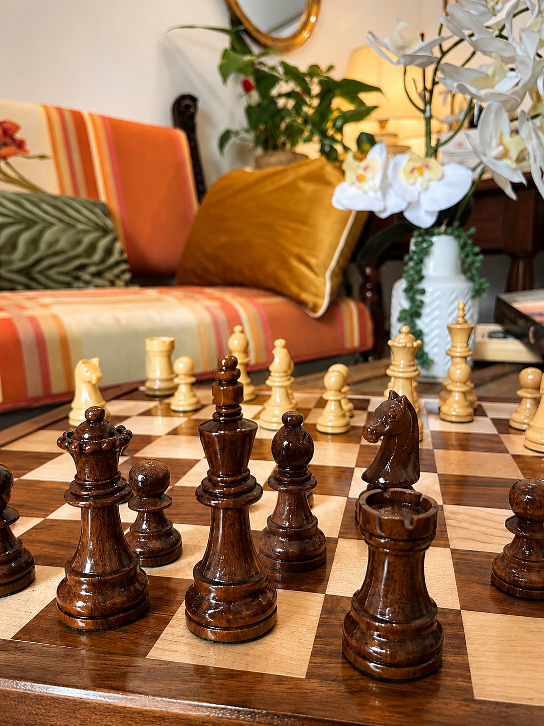 Pièces d'échecs en bois sombre et clair disposées sur un échiquier, avec un canapé orangé, un coussin moutarde et des orchidées blanches en arrière-plan.
