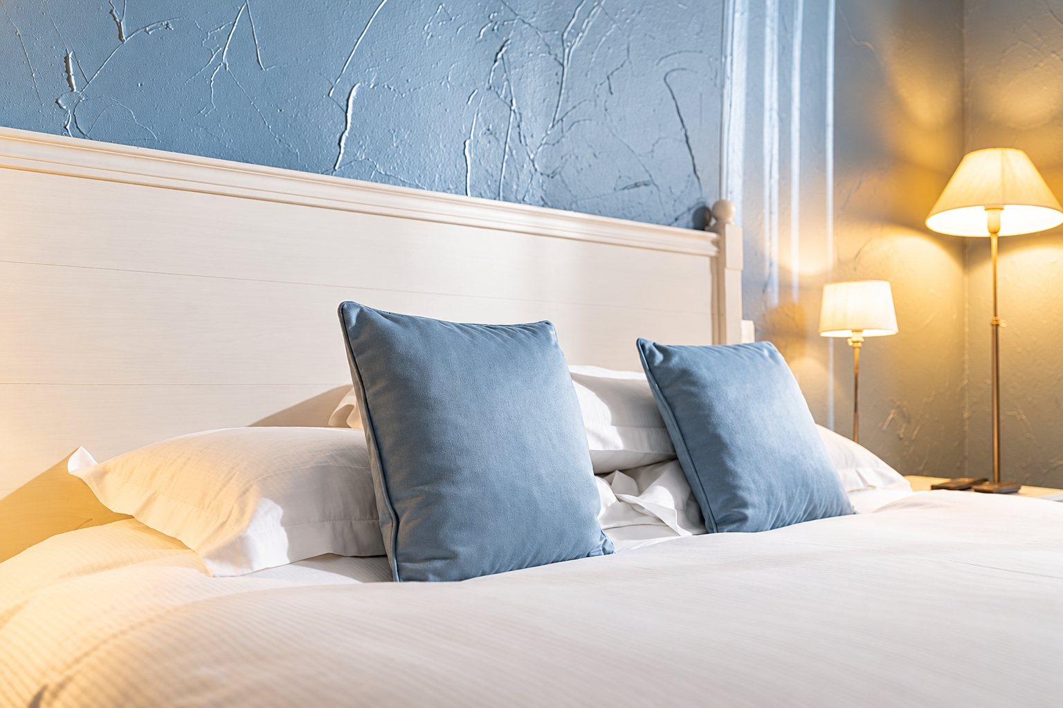 Un lit habillé de linge blanc et de deux coussins bleus devant une tête de lit blanche et un mur bleu texturé, avec des lampes allumées en arrière-plan.