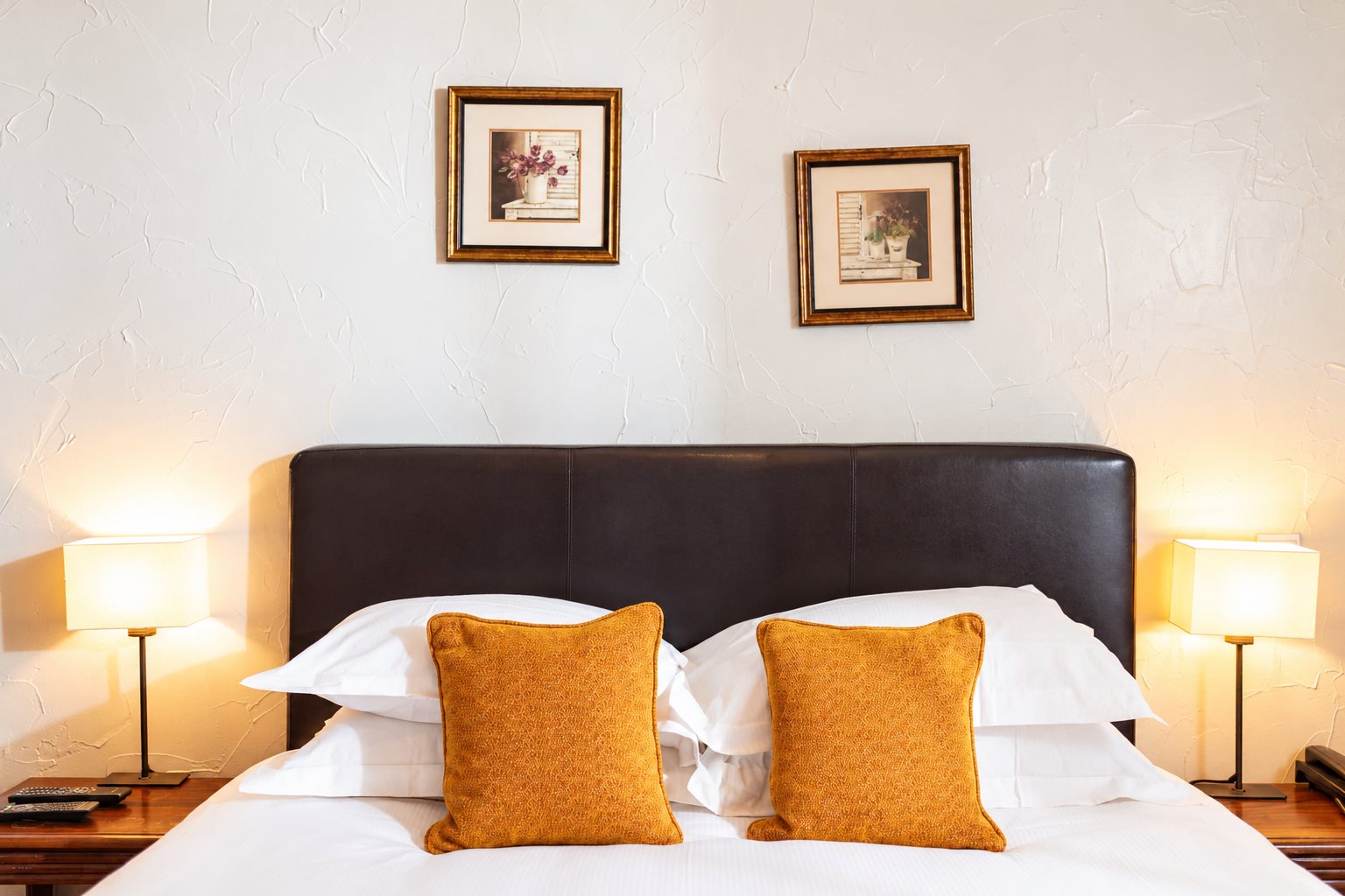 Chambre d'hôtel avec un lit double à tête de lit en cuir brun, deux coussins orange, deux lampes de chevet allumées et deux cadres au mur blanc.