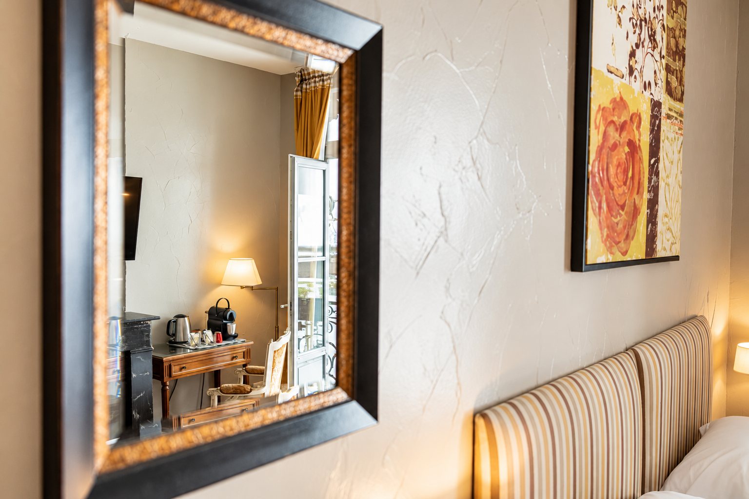 Un miroir au cadre sombre et doré reflète un petit bureau avec bouilloire et machine à café, à côté d'une tête de lit rayée et d'un tableau coloré.