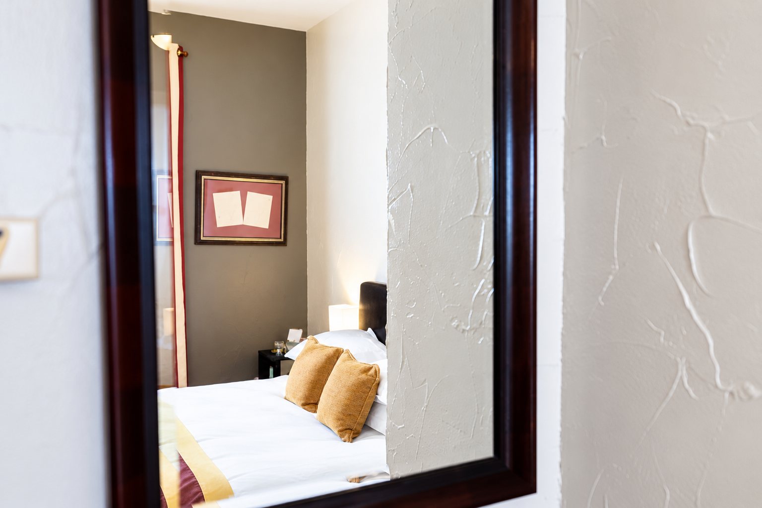 Un miroir au cadre en bois sombre reflète un lit blanc avec des coussins orangés, un cadre rose au mur et un rideau, sur un mur blanc texturé.