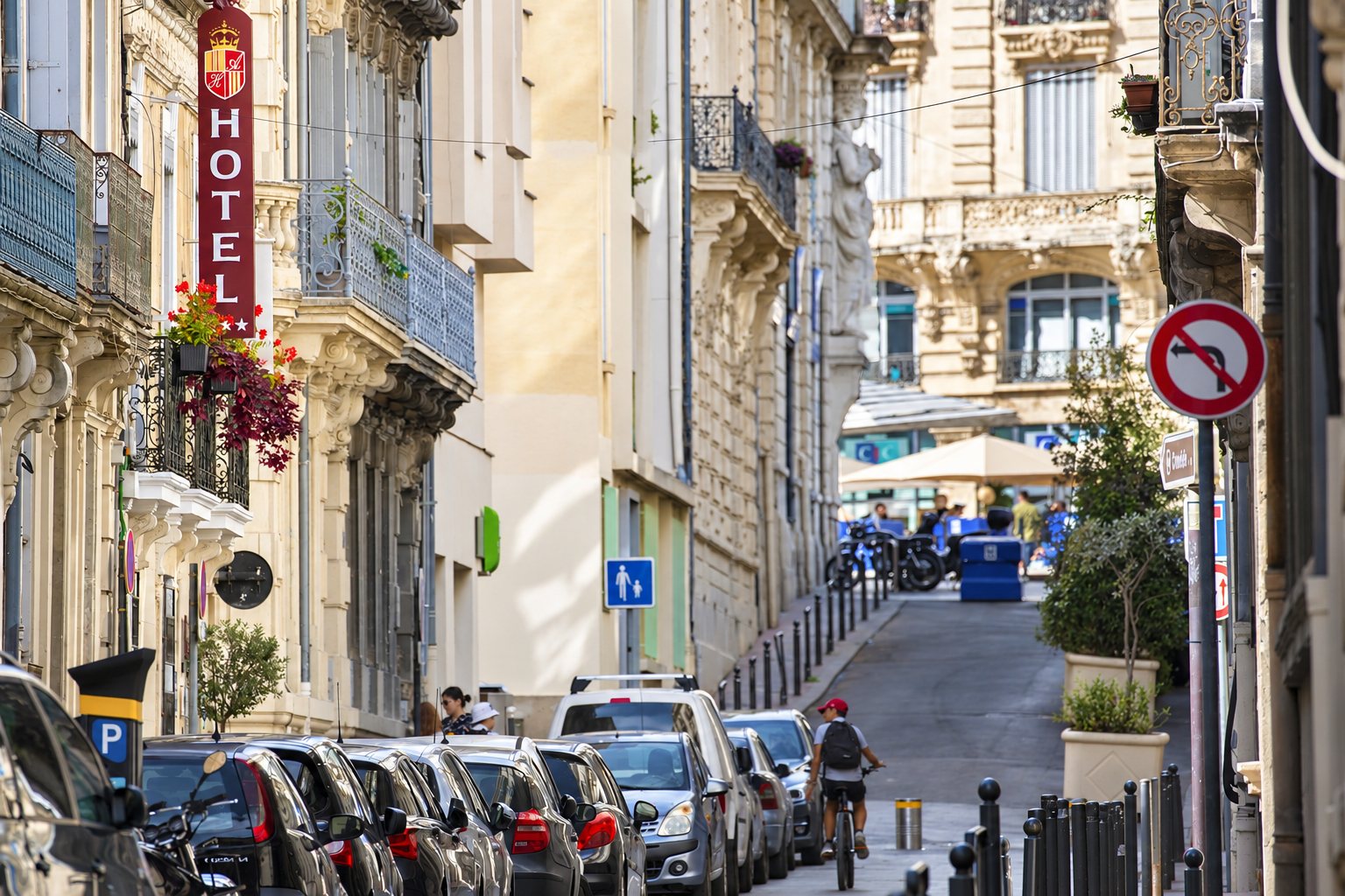 Une rue étroite bordée d'immeubles anciens, avec une enseigne verticale d'hôtel, des voitures garées et un cycliste qui descend la pente.