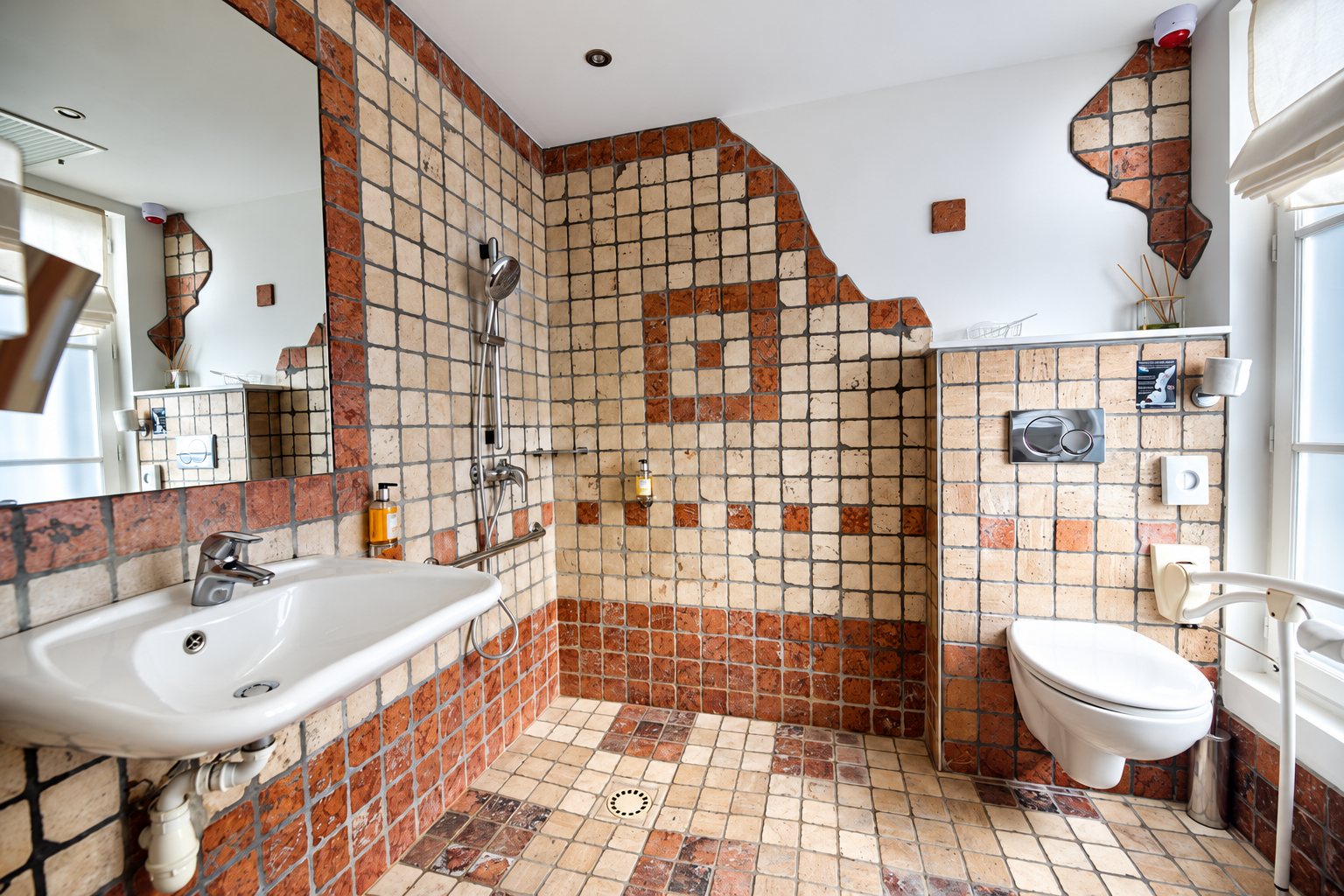Salle de bain aux murs et au sol en petits carreaux beiges et orangés, avec lavabo suspendu, douche à l'italienne, WC suspendu et barres d'appui.