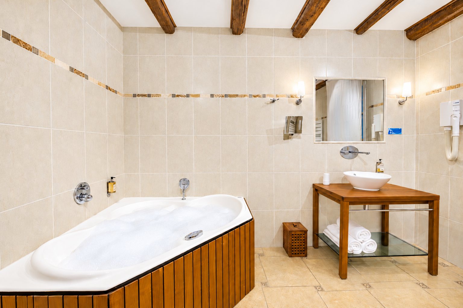 Salle de bain aux murs carrelés beige avec une grande baignoire d'angle remplie de mousse, une vasque blanche posée sur un meuble en bois et des poutres apparentes au plafond.