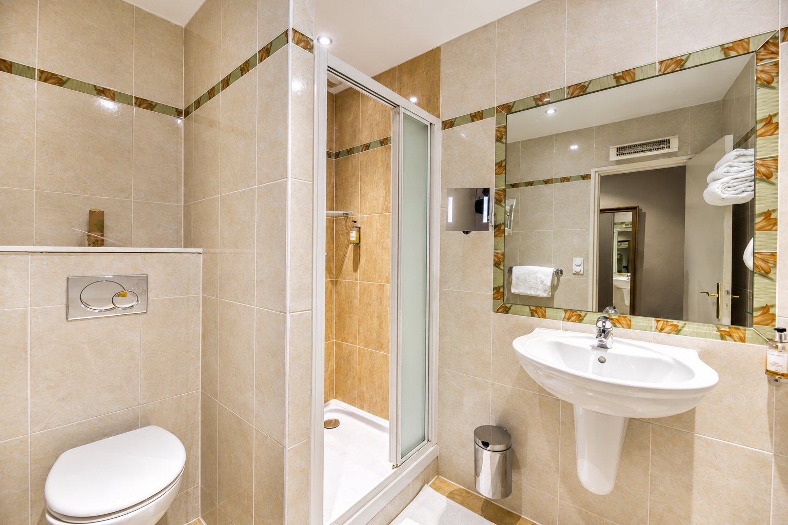 Salle de bain carrelée de beige avec une cabine de douche, un WC suspendu, un lavabo sur colonne et un miroir à bordure fleurie.