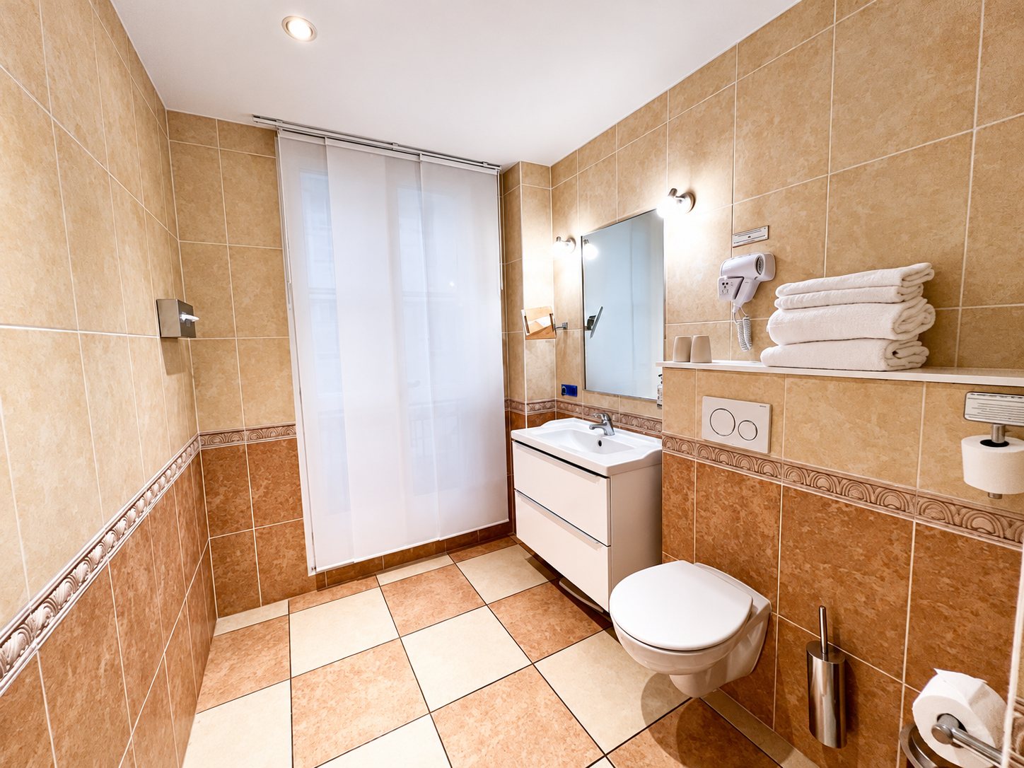 Salle de bain carrelée de beige avec un meuble vasque blanc, un WC suspendu, un miroir, des serviettes blanches empilées et un store à panneaux.