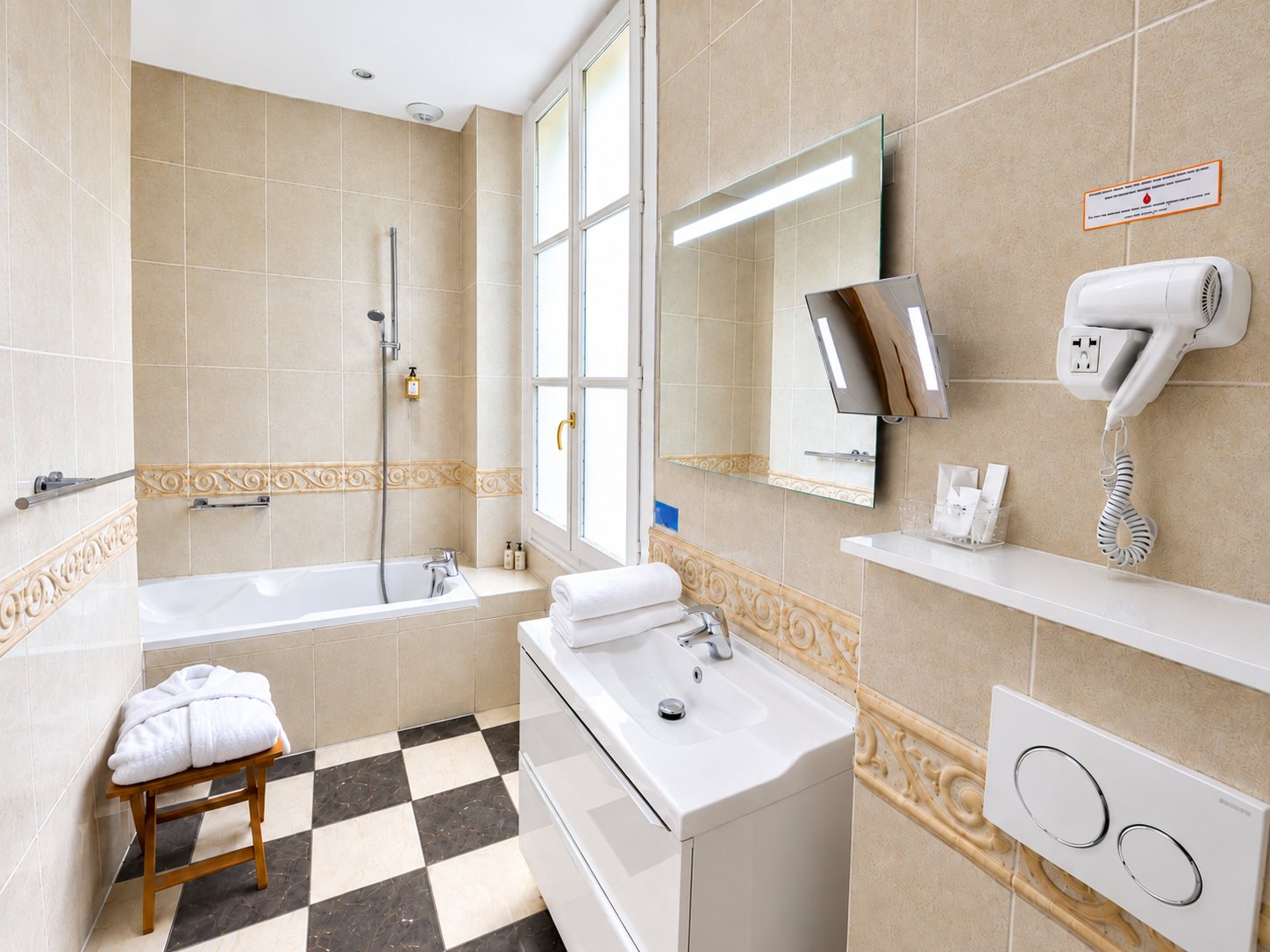 Salle de bain d'hôtel aux carreaux beiges avec baignoire, vasque blanche, miroir lumineux, sèche-cheveux mural et sol en damier noir et blanc.