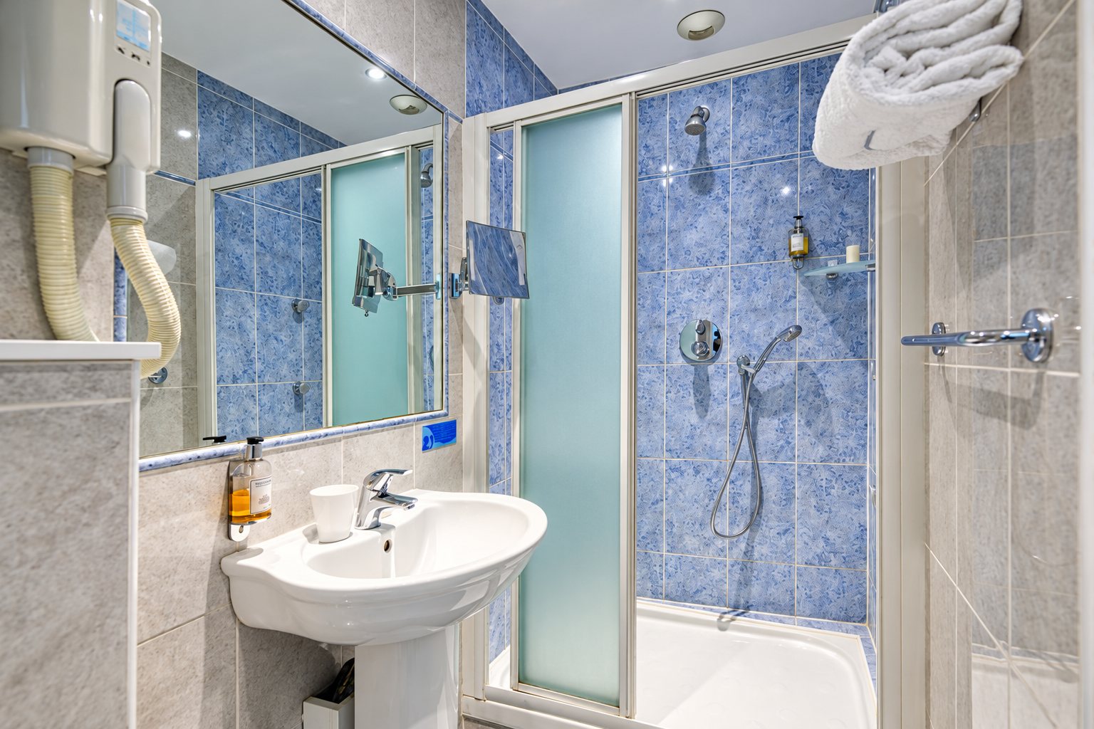 Salle de bain avec une cabine de douche à parois vitrées, un lavabo sur pied, un grand miroir, un sèche-cheveux mural et un carrelage bleu et gris.