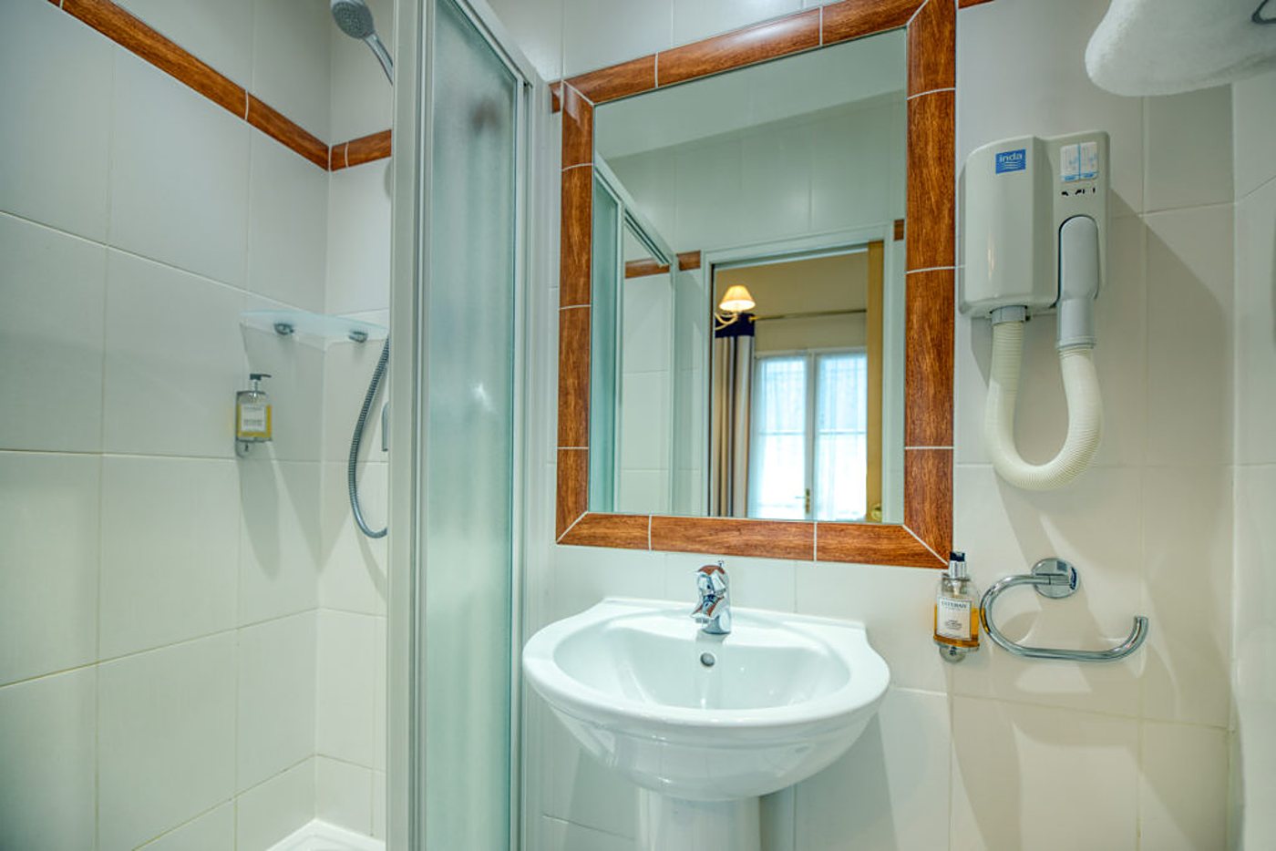 Salle de bain carrelée de blanc avec une cabine de douche, un lavabo sur colonne, un miroir à cadre orange et un sèche-cheveux mural.