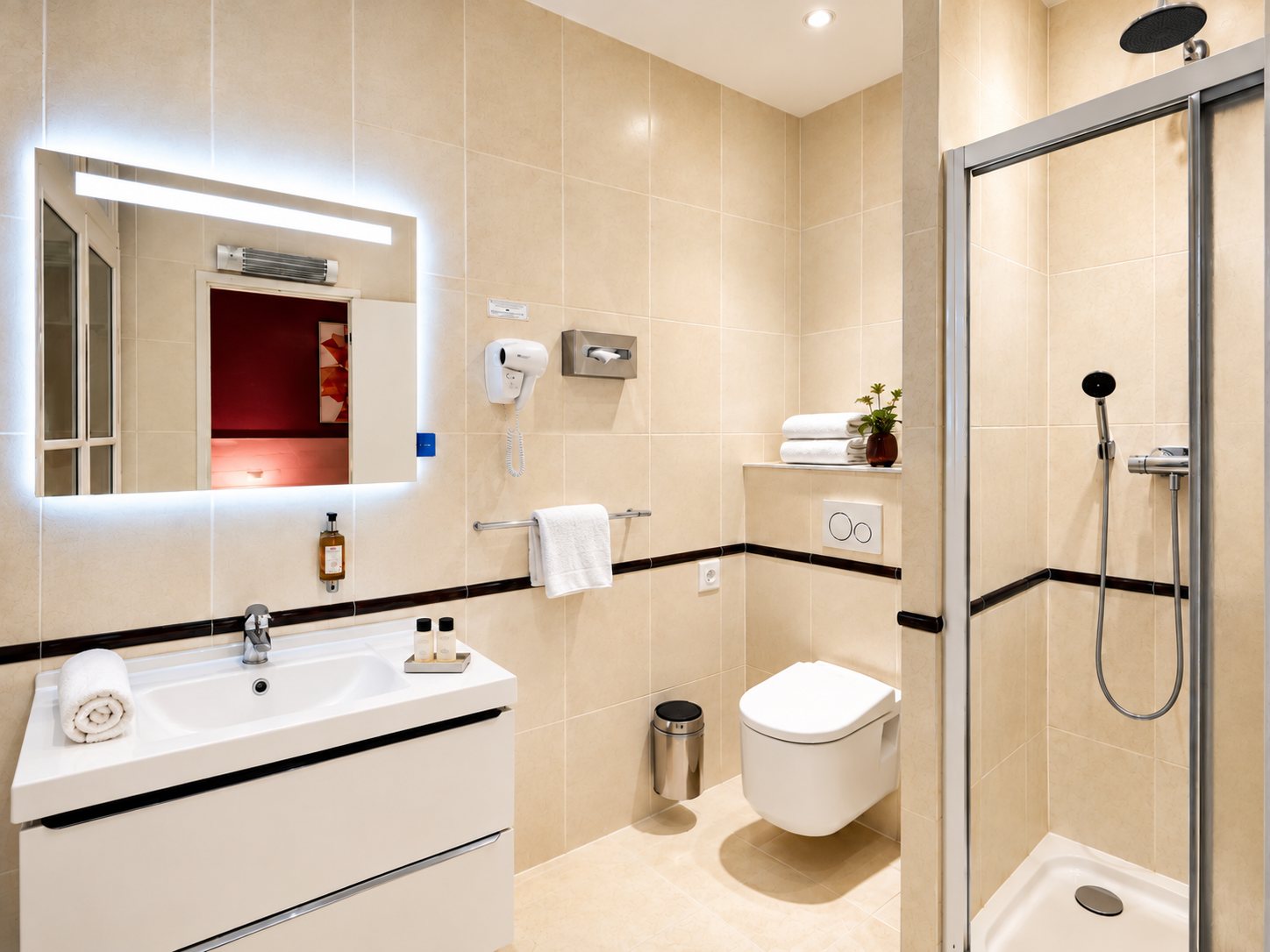 Salle de bain d'hôtel aux carreaux beiges avec vasque blanche, miroir rétroéclairé, WC suspendu et douche à l'italienne.