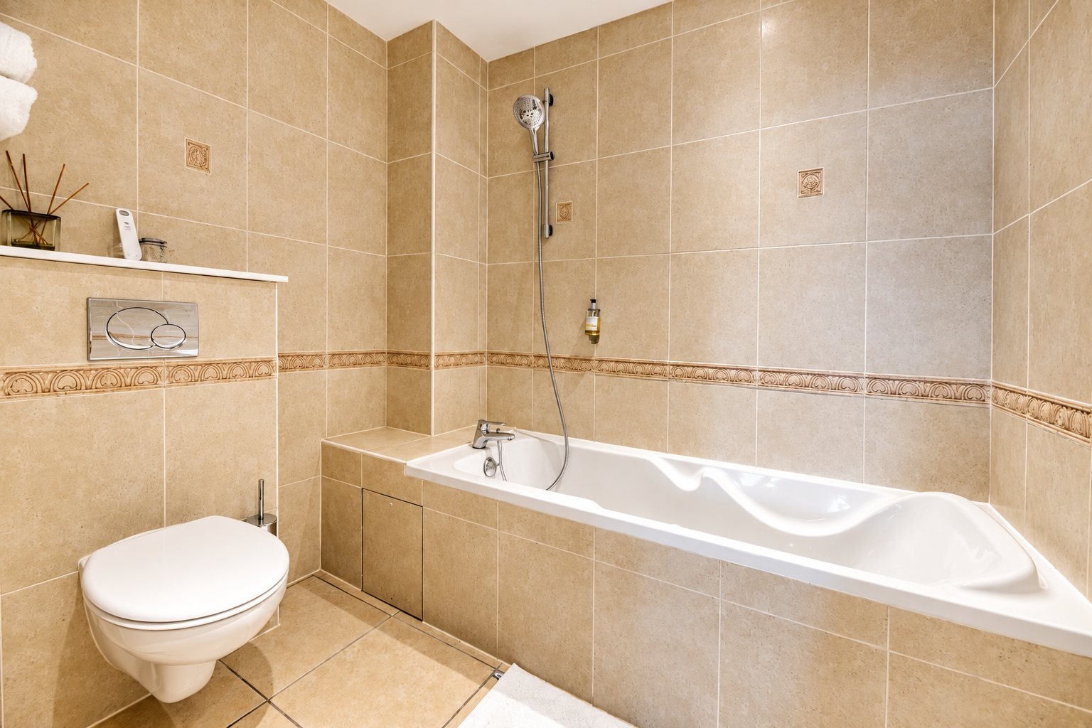 Salle de bains aux murs carrelés beige avec une baignoire blanche, une douchette murale, un WC suspendu et une frise décorative.