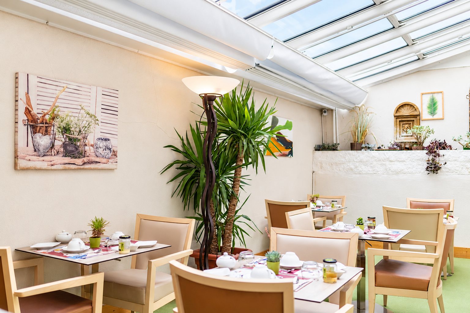 Salle de petit-déjeuner sous une verrière, avec des tables dressées de tasses et de pots de confiture, des chaises beiges, un grand palmier et un lampadaire.