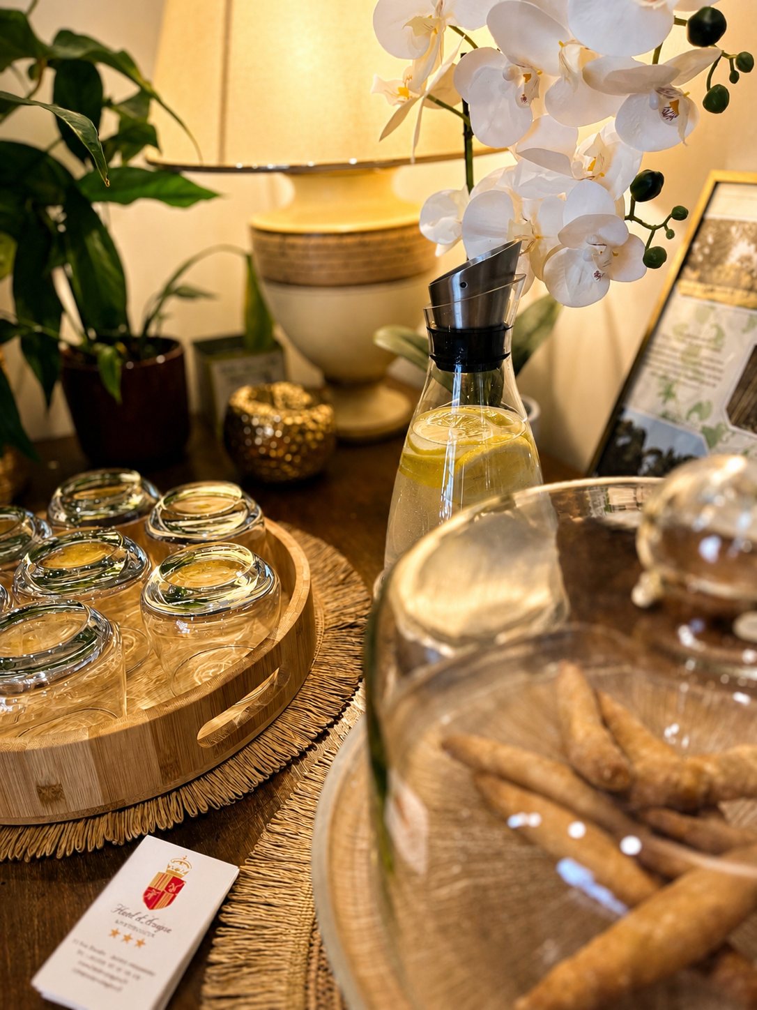 Sur une table en bois, un plateau de verres retournés, une carafe d'eau au citron, une orchidée blanche, des biscuits sous cloche et une carte de visite d'hôtel.